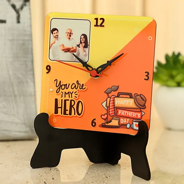 A square, personalized table clock for dad with a photo of a family, the text "You are my HERO", and Father's Day themed illustrations, resting on a black stand. The clock face is yellow and orange.